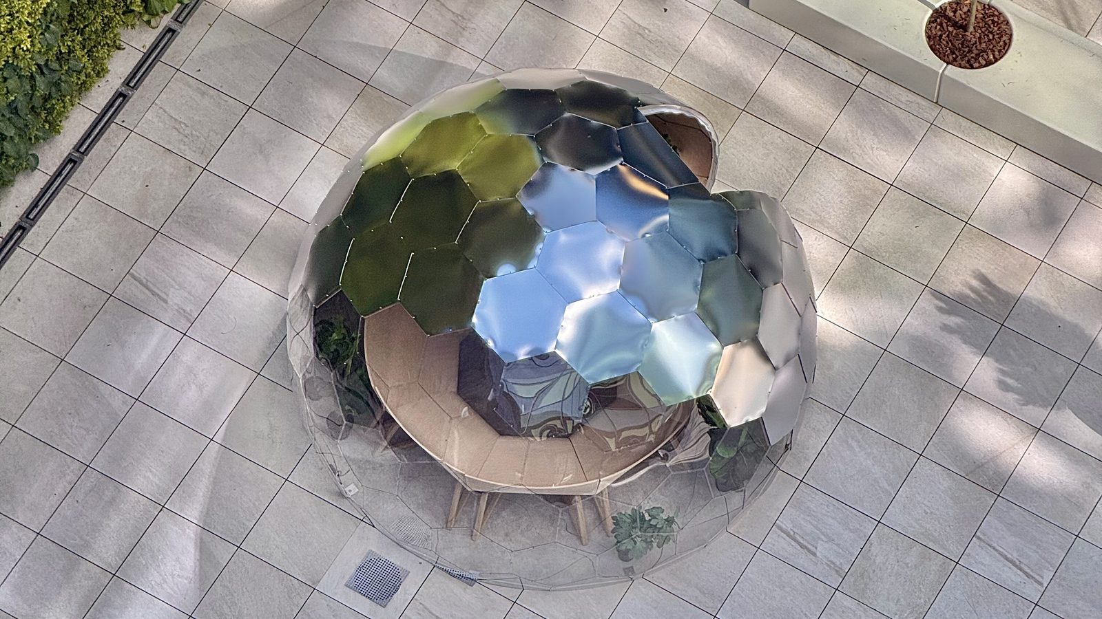 Aerial view of a satin-mirror Hypedome in a landscaped courtyard, its facets reflecting the green wall and sky