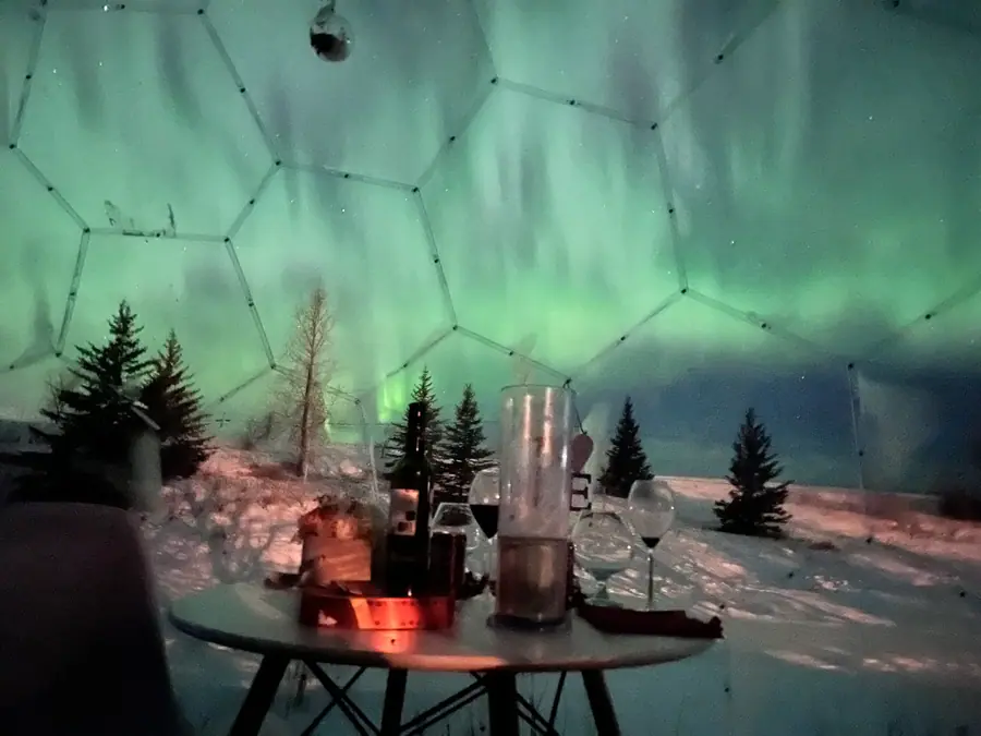 Candlelit table inside a dome with an aurora scene over snowy pines
