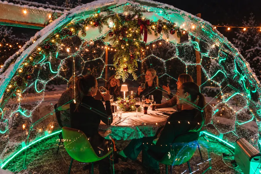 Friends laughing and toasting at a candlelit festive dinner inside a snow-dusted Hypedome with garlands and green LED lighting