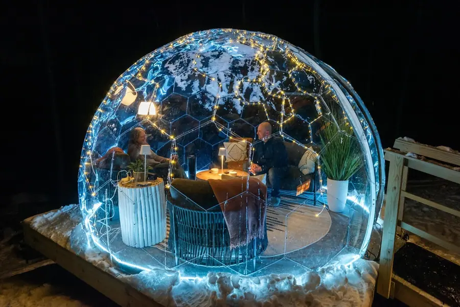 Glowing transparent dome on a snowy mountain deck at night with two guests inside