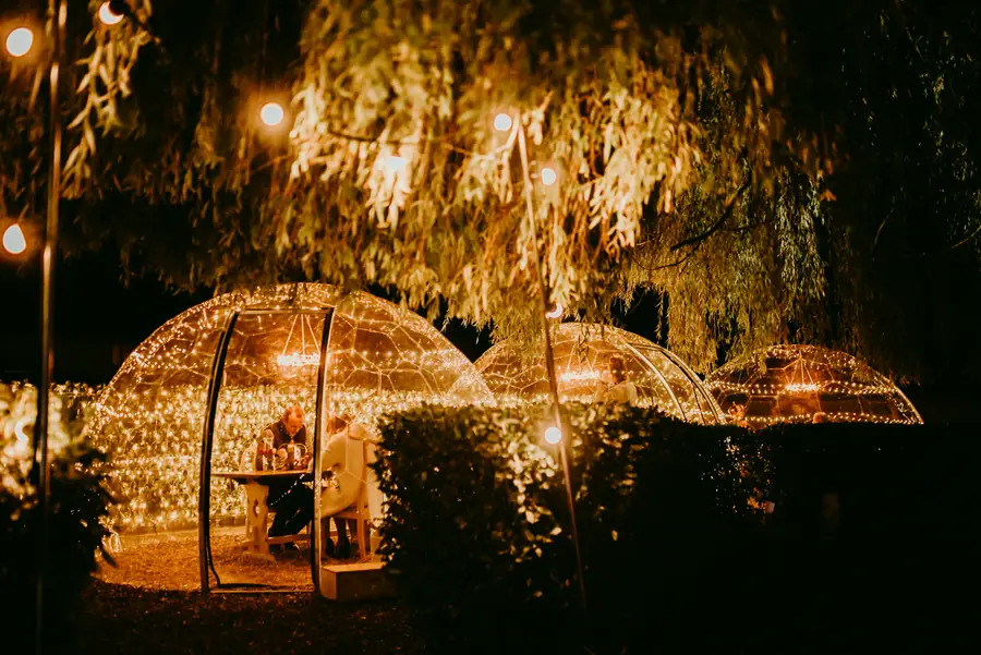 Fairy-lit Hypedome dining domes in a restaurant garden under weeping willow trees, with a couple dining inside