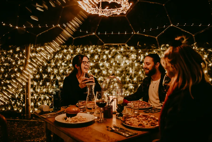 Four friends laughing over pizza and wine inside a fairy-lit dome at The Willow Tree