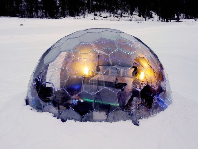 A Hypedome glowing with warm light and fairy lights, half-covered in snow on a frozen lake at dusk